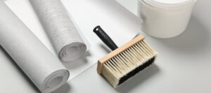 différents rouleaux de papier-peint, pot de colle à tapisserie et pinceau à tapisser sur fond gris clair