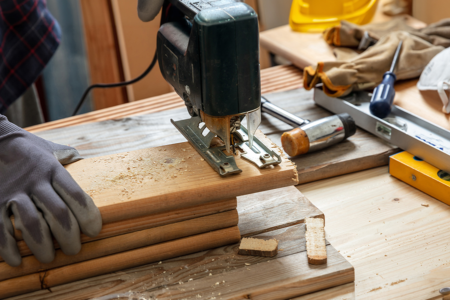 gros plan sur les mains du menuisier qui manipulent une scie sauteuse pour couper des planches de bois, des outils sont posés sur son établi dans l'atelier de menuiserie
